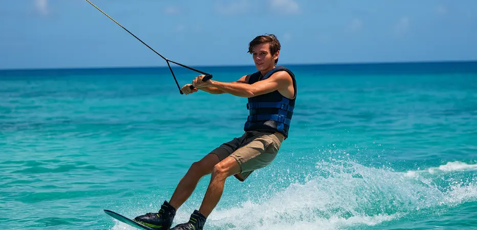 A young man wakeboarding across turquoise Caribbean waters, with a sunny sky and tropical shoreline in the background.
