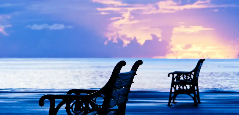 Two benches sit on the boardwalk in Bridgetown, Barbados with a purple and yellow sunset in the background.