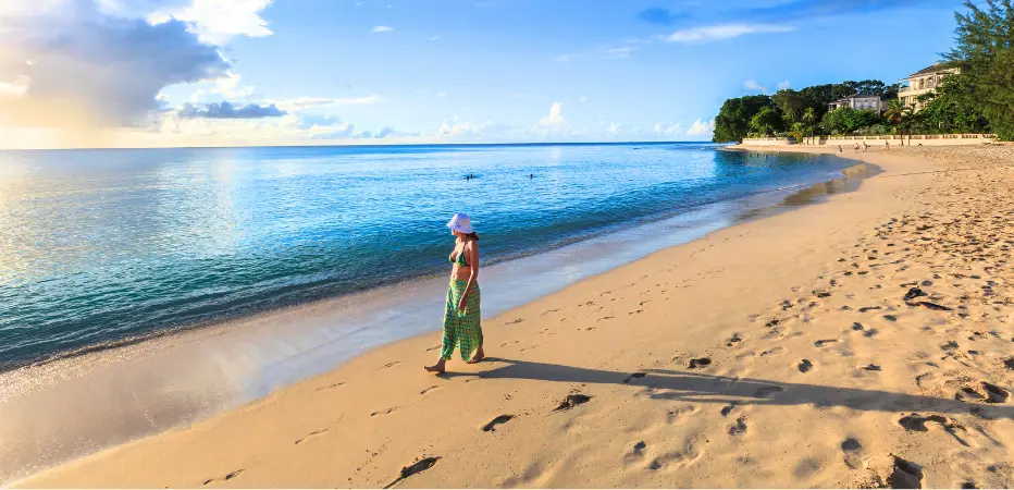 A lady strolls along the beach, looking towards the sun over turquoise waters
